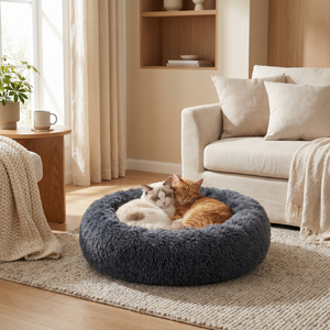 Two cats snuggling in a cozy, plush gray pet bed in a warm living room setting.